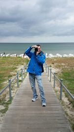 Full length of man photographing on boardwalk at beach against cloudy sky