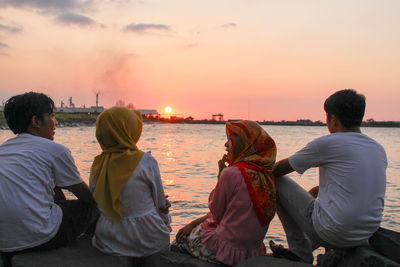 Rear view of friends sitting by lake against sky during sunset