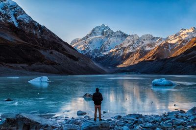 Rear view of man standing by lake against snowcapped mountain
