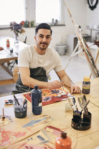 Portrait of confident young male student painting while sitting at table in art class