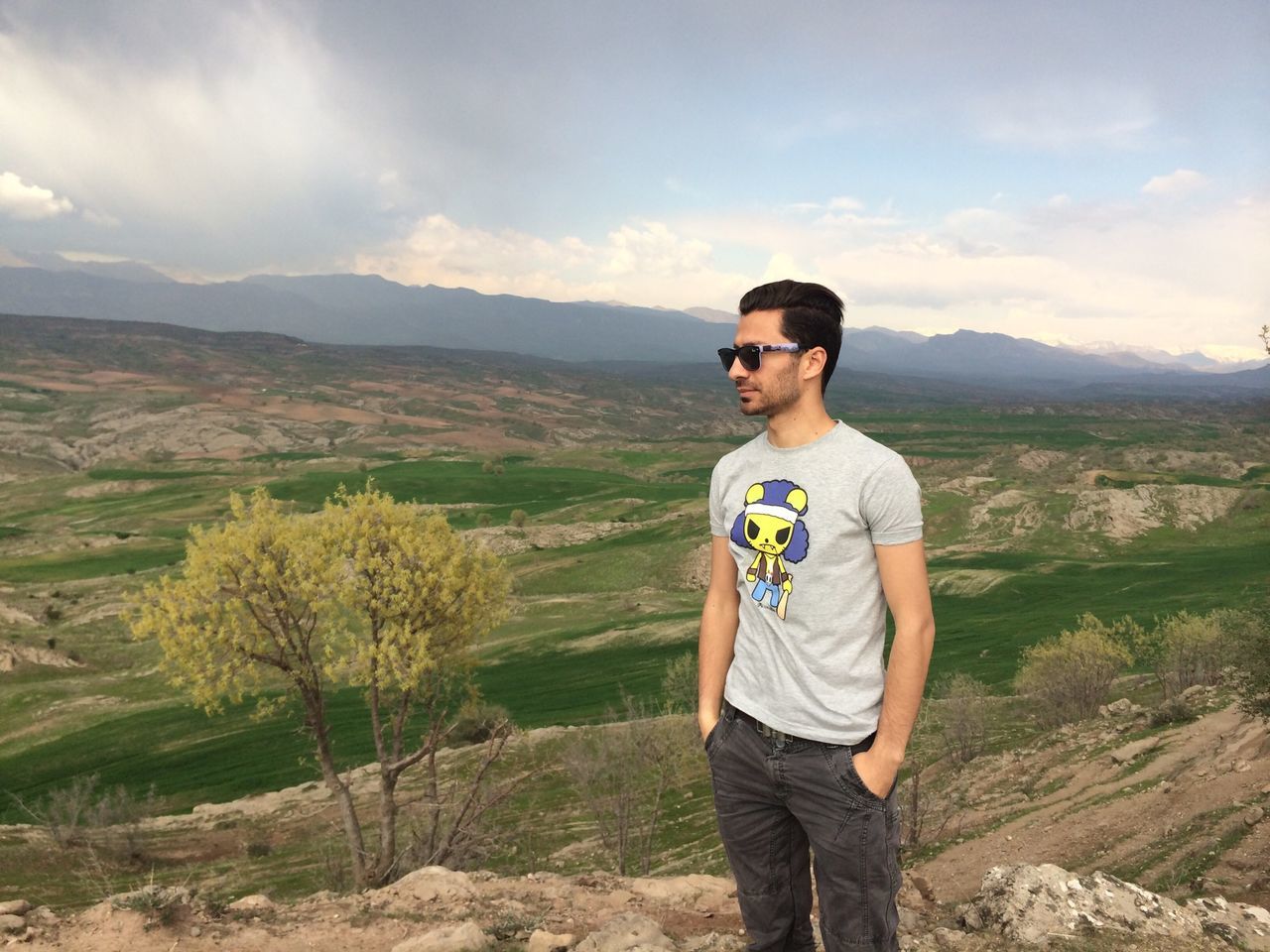 casual clothing, mountain, young adult, lifestyles, person, sky, leisure activity, young men, landscape, standing, portrait, looking at camera, mountain range, sunglasses, front view, smiling, field, three quarter length