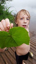 High angle portrait of shirtless boy showing leaf while standing at riverbank