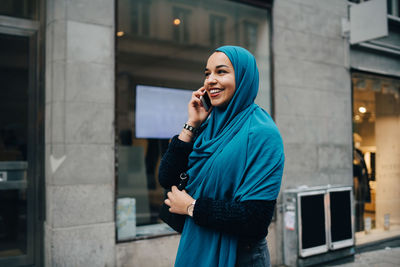 Happy young muslim woman talking on mobile phone walking building in city