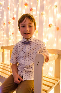 Portrait of boy sitting on bench against decorative curtain