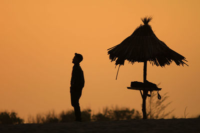 Silhouette man standing by thatched roof against orange sky