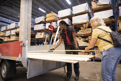 Smiling female customer loading plank with saleswoman in trailer at hardware store