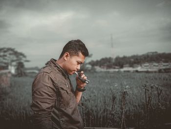 Side view of young man smoking cigarette on field against sky