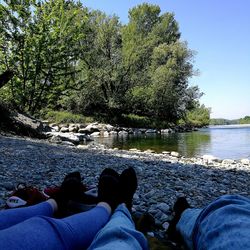 Low section of men sitting on rock by river against sky