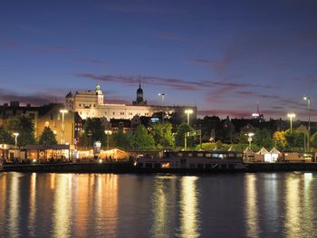 Illuminated buildings by river against sky at sunset