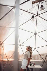 Side view of young woman standing at cafe during sunset