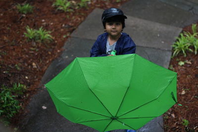 High angle portrait of boy holding green umbrella while standing on wet pathway