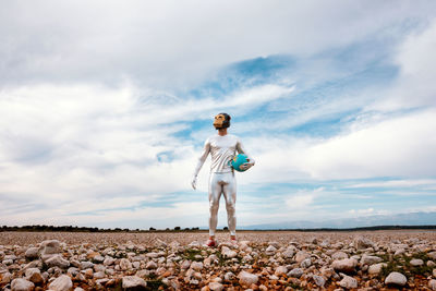 Anonymous guy in silver latex suit with geometric monkey mask looking away and holding globe in nature