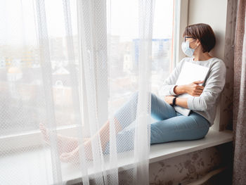 Woman in medical mask had lost her job. she sits on window sill with closed laptop. 