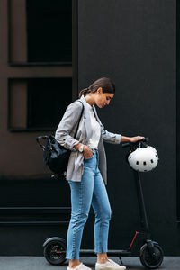 Full length shot of young businesswoman standing at a building with electric push scooter 	
