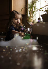 Playful girl filling water while sitting amidst soap sud in kitchen sink at home