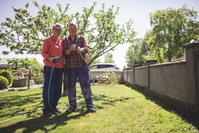 Full length portrait of senior couple holding hammer and garden hose while standing at yard