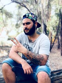 Portrait of young man gesturing while sitting on log in forest