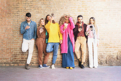 Full length of friends with smart phones standing against brick wall