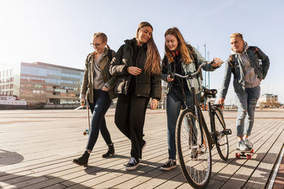 Teenage talking to friend holding bicycle while boy skateboarding at city