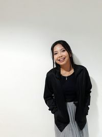 Portrait of smiling young woman with hands in pockets standing against white background