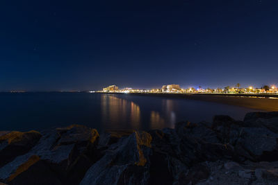 Illuminated city by sea against sky at night