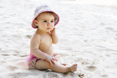 Cute little baby girl in pink swimming trunks and panama sitting on sand at beach.