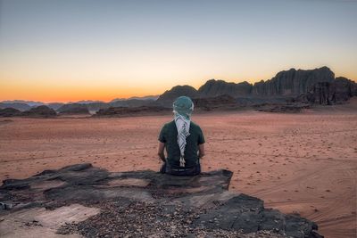 Man sitting on rock in desert against sky during sunset