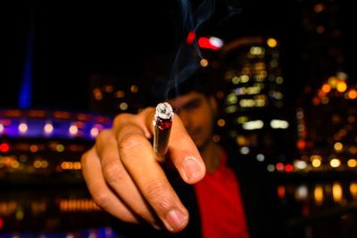 Man showing burning cigarette while standing in illuminated city at night