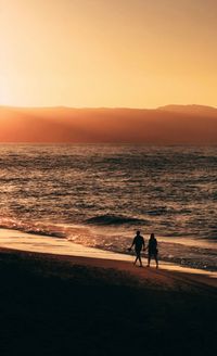 Silhouette couple walking on beach against sky during sunset