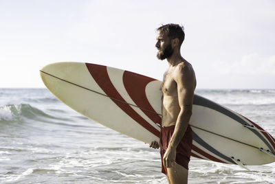 Young man with surfboard at beach against sky