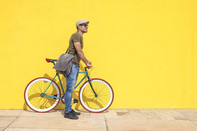 Side view of man with bicycle standing on footpath against yellow wall