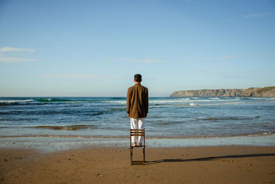 Back view of anonymous male model in stylish suit standing on chair on beach and admiring waving sea