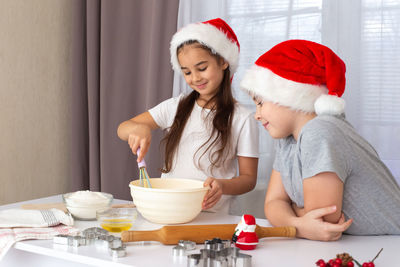 Happy children, brother and sister, in red caps, prepare christmas cookies in the kitchen