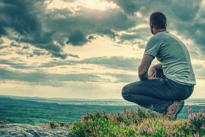 Tall man sits on a hillside in heather and admires the tops of hills touch in the clouds.