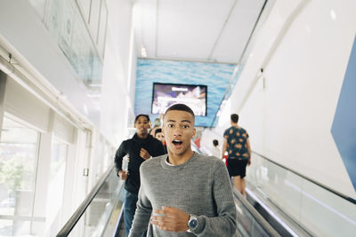Portrait of shocked teenage boy moving down on escalator with friend in background at shopping mall