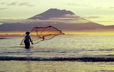 Man throwing fishing net on sea against mountain during sunset