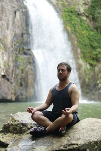 Young man sitting in lotus position on rock against waterfall