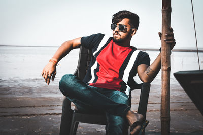 Young man wearing sunglasses sitting by sea against sky