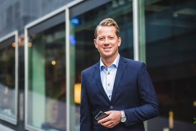 Portrait of smiling mature businessman with smart phone standing against office building