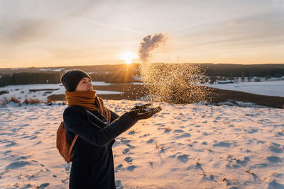 A woman in a snow-covered field tosses snow with her hands against the backdrop of a sunset