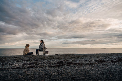 Friends talking while resting on sea shore at beach against cloudy sky