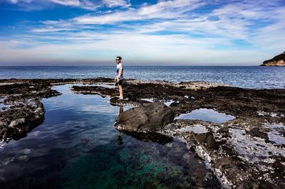 Young man standing on rock by sea against sky