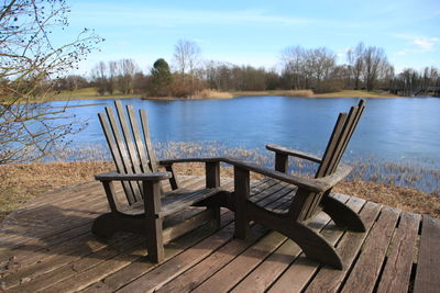 Empty chairs and table by lake against sky