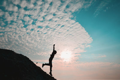 Silhouette boy with arms raised on rock against sky