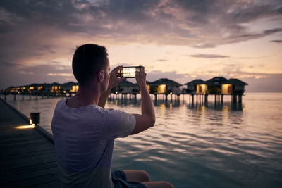 Young man with mobile phone photographing seascape with water bungalows at sunset.