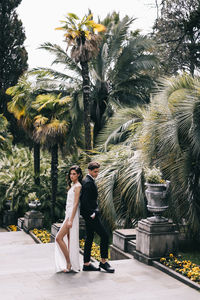 Happy lovers the bride and groom in wedding outfits walk among plants and palm trees in the old park