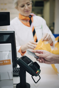 Cropped hand of male customer using contactless payment through credit card against pharmacist at checkout in store