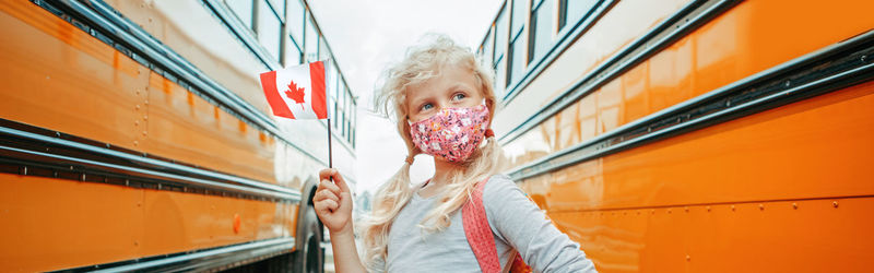 Close-up of cute girl holding canadian flag standing amidst school bus