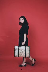 Portrait of young woman with luggage standing against red background