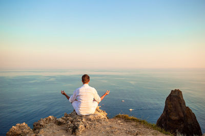 Rear view of woman sitting on rock looking at sea against sky